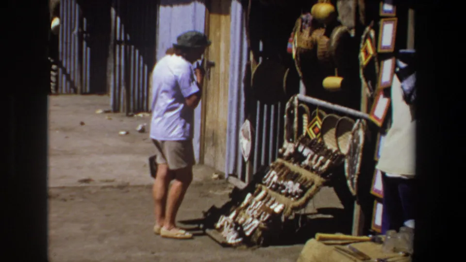 The tourist pauses beside the craft display, with woven items propped against a corrugated wall and additional wares hanging near the doorway.