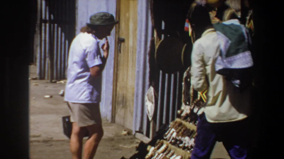 In the final frame, the tourist stands near the craft stall while the vendor passes close to camera beside baskets, hanging hats, and framed items.