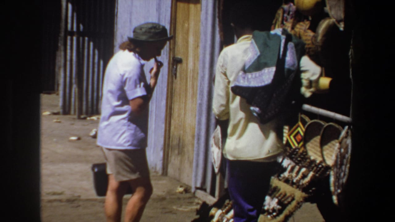In 1969 Pretoria, a woman in a light shirt and brimmed hat stands beside a market stall as a vendor faces displayed baskets and craft goods.