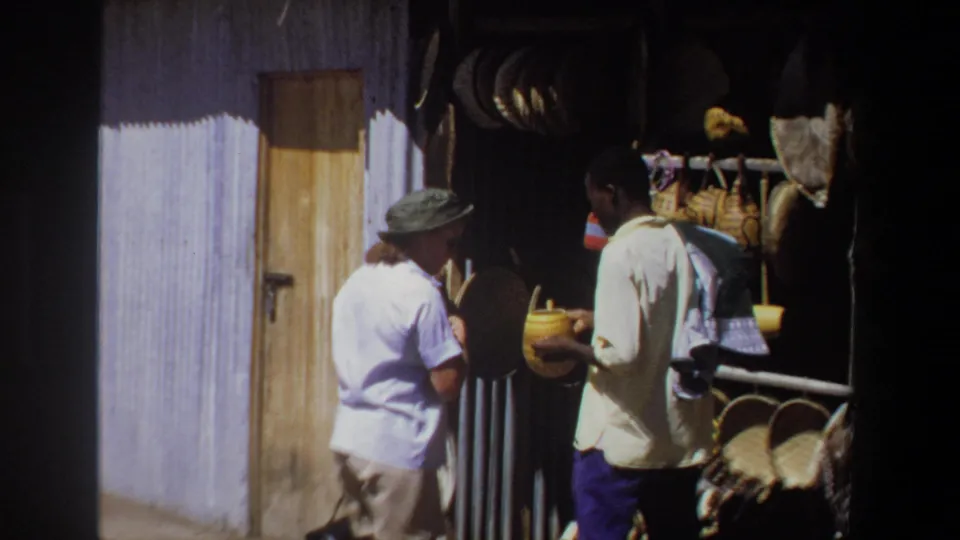 In the Pretoria market scene, the vendor presents or holds a yellow container as the woman stands close to the craft display.