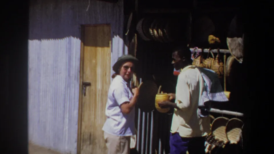 The woman briefly looks toward the camera during the 1969 Pretoria market exchange as the vendor remains beside the baskets and hanging wares.
