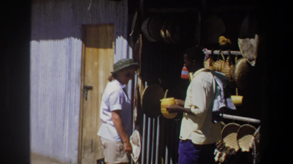 The Pretoria market exchange continues as the woman stands near the vendor, yellow container, baskets, and craft items in bright daylight.