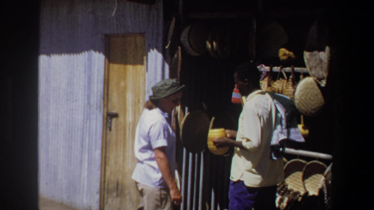 In 1969 Pretoria, a woman in light clothing stands near a vendor holding a yellow handmade object beside hanging baskets and crafts.
