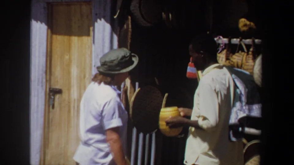 The Pretoria market scene shows the vendor holding an object while the woman looks toward a display of woven baskets and crafts.