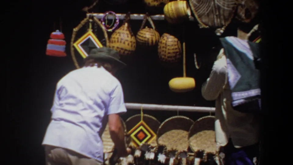 At the craft stall in Pretoria, the woman leans toward displayed goods while woven baskets and colorful hanging objects fill the shaded background.