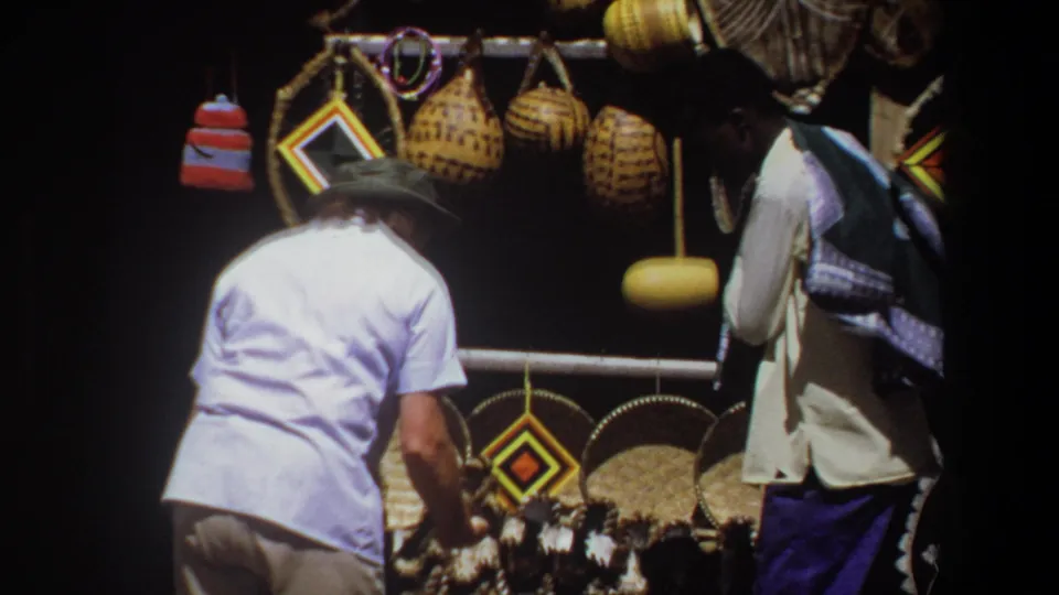 In Pretoria, South Africa in 1969, a white-hatted man browses woven baskets and geometric craft items at a shaded outdoor stall.