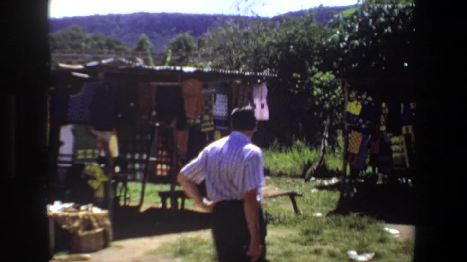 In a Pretoria outdoor market setting, a man stands with his back to camera near stalls, greenery, and distant hills.