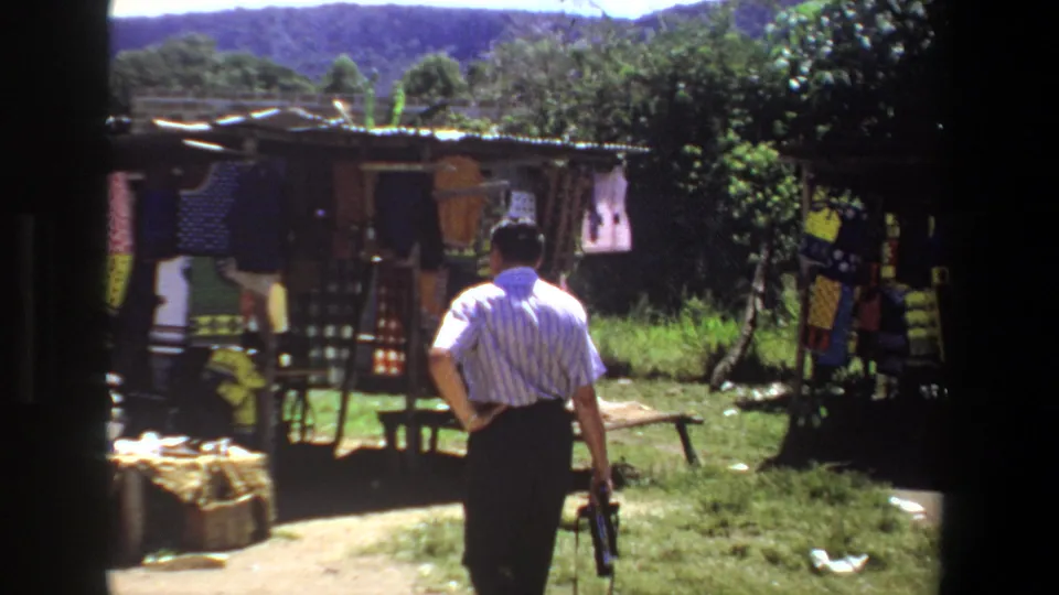 A 1969 Pretoria market scene shows a man walking past craft displays, grass, trees, and low hills in a handheld home-movie frame.