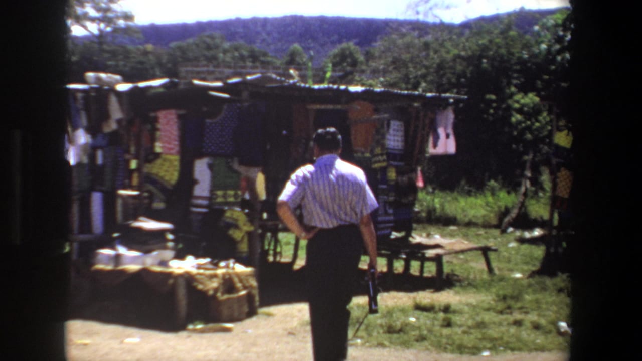 In Pretoria, South Africa, 1969, a man in a striped shirt approaches a small outdoor stall displaying hanging fabrics and folded cloth.