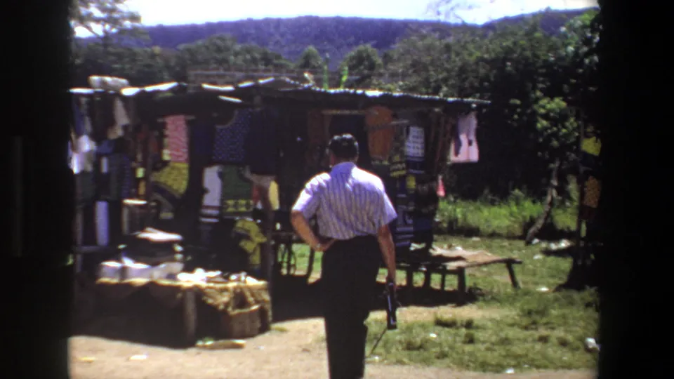 In Pretoria, South Africa, 1969, a man in a striped shirt approaches a small outdoor stall displaying hanging fabrics and folded cloth.