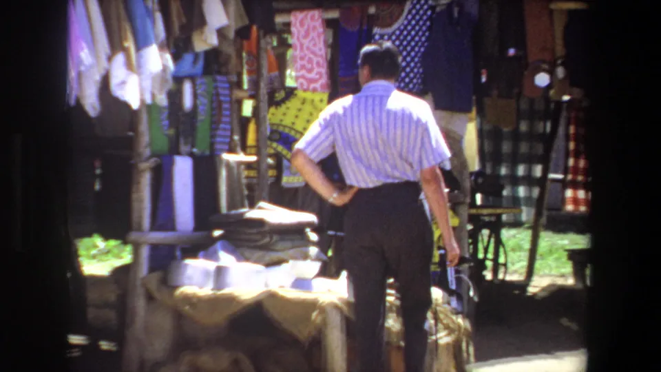 A closer 1969 view shows the man pausing before colorful fabrics and stacked textiles at the small outdoor stall in Pretoria.