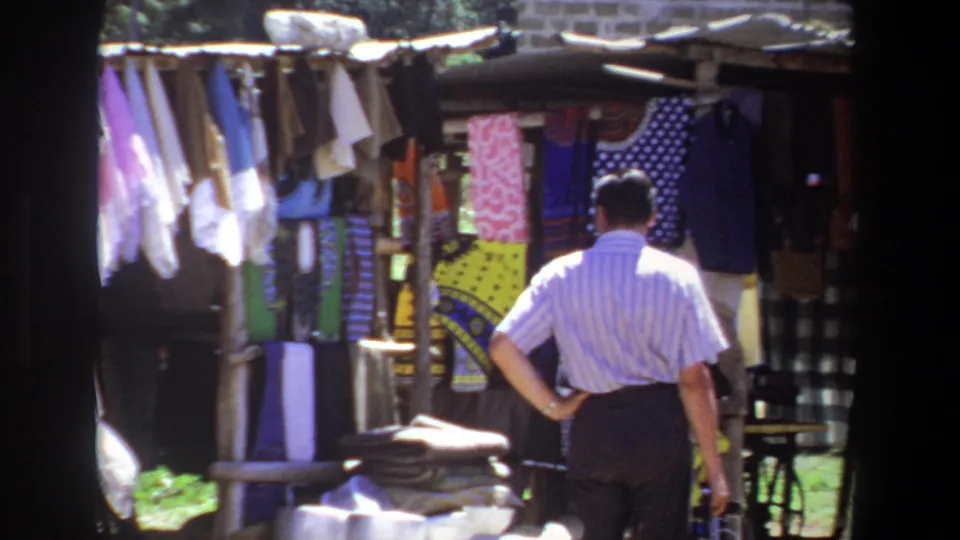 At the end of the clip, the man stands near a table of folded cloth while facing hanging textiles at the Pretoria outdoor stall.