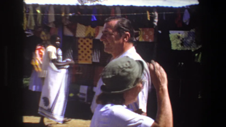 At the Pretoria market, two adult men and the woman in a green hat gather near a stall displaying hanging fabrics and clothing.