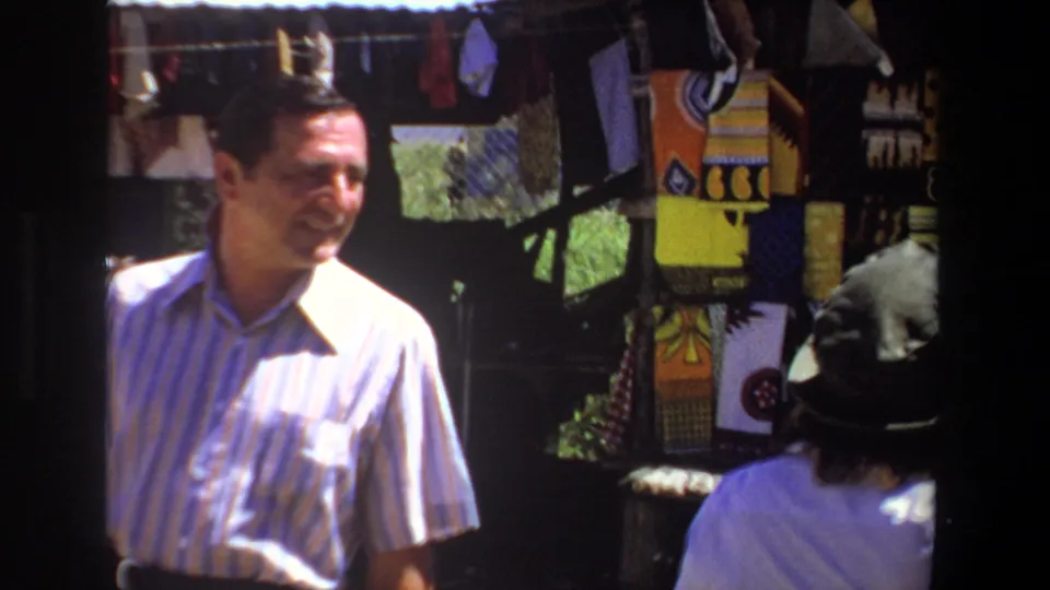 A man in a striped shirt smiles beside colorful patterned textiles while the woman in a green hat is partly visible at the Pretoria market stall.