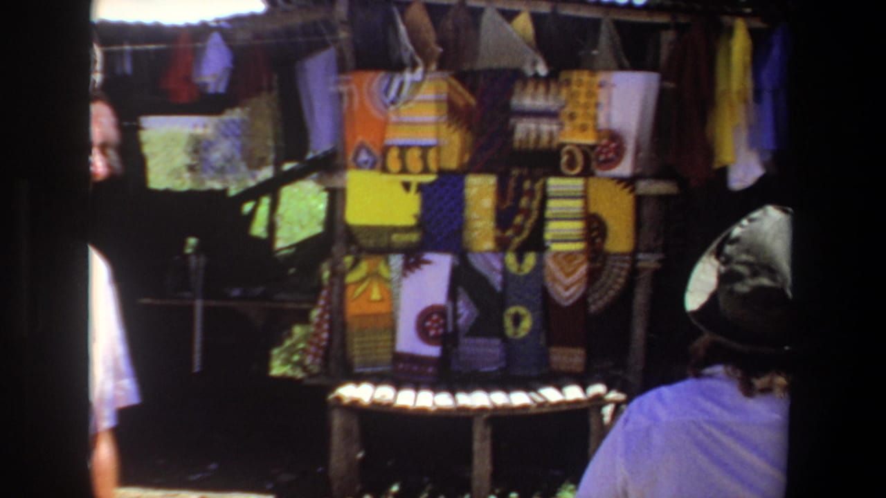 1969 Pretoria home-movie frame showing a woman in a brimmed hat looking toward colorful textiles at an outdoor market stall.