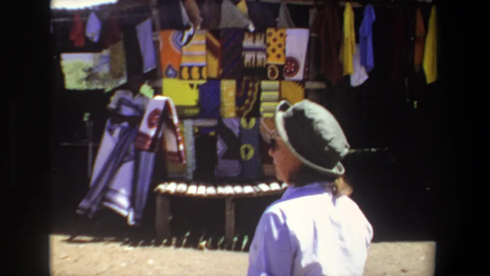 1969 Pretoria home-movie frame of a woman near an outdoor textile display, with colorful fabrics filling the stall behind her.
