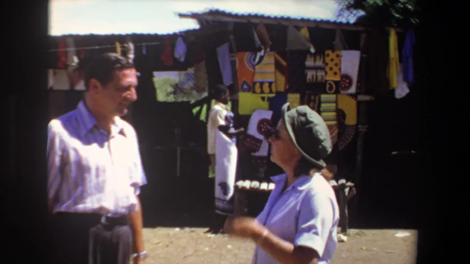 1969 Pretoria market frame showing a man in a striped shirt and a woman in a brimmed hat beside a colorful textile stall.
