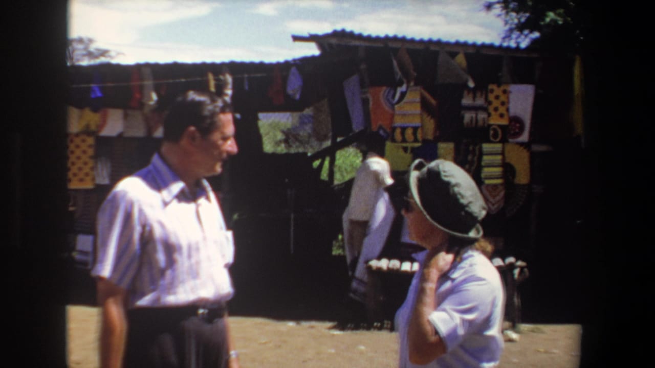 In 1969 Pretoria home-movie footage, two adult visitors appear near an outdoor market stall displaying colorful textiles.