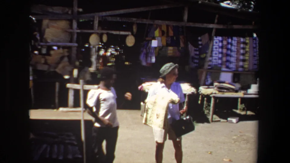 The home-movie frame shows a woman carrying a folded rug through a sunlit Pretoria market area with textile stalls behind her.