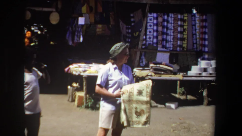 A woman in 1969 Pretoria market footage holds a patterned rug while standing before shaded displays of textiles and goods.