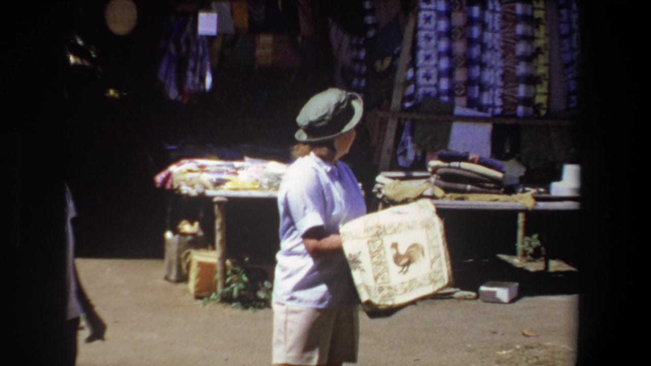 In 1969 Pretoria home-movie footage, a woman in a light shirt and hat holds a decorative rooster tapestry near hanging fabrics at a roadside stall.