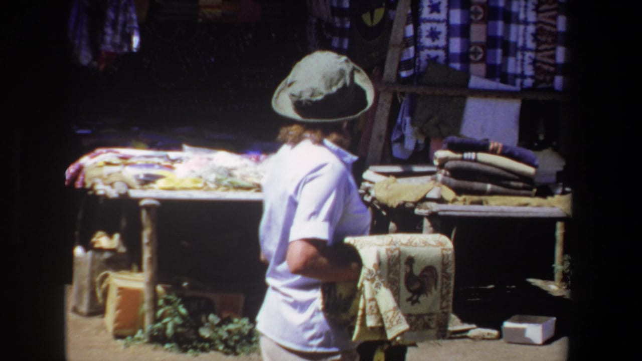 Woman turns at the market stall while holding the rooster tapestry and looking toward hanging fabrics.