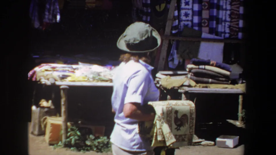 The woman stands side-on in the Pretoria market scene, holding the rooster tapestry near a stall with patterned textiles and folded goods.