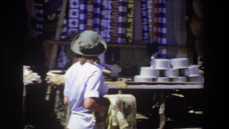 The camera follows the woman from behind as she carries the tapestry past hanging fabrics and stacked white bowls at the outdoor market.