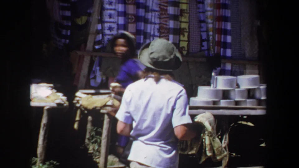 At the end of the clip, the woman approaches the stall with the tapestry while another person is partly visible behind the market display.