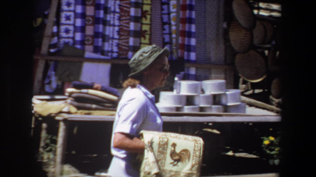 In 1969 Pretoria footage, a woman in a brimmed hat walks through an outdoor market holding a rooster-patterned textile near baskets and stacked goods.