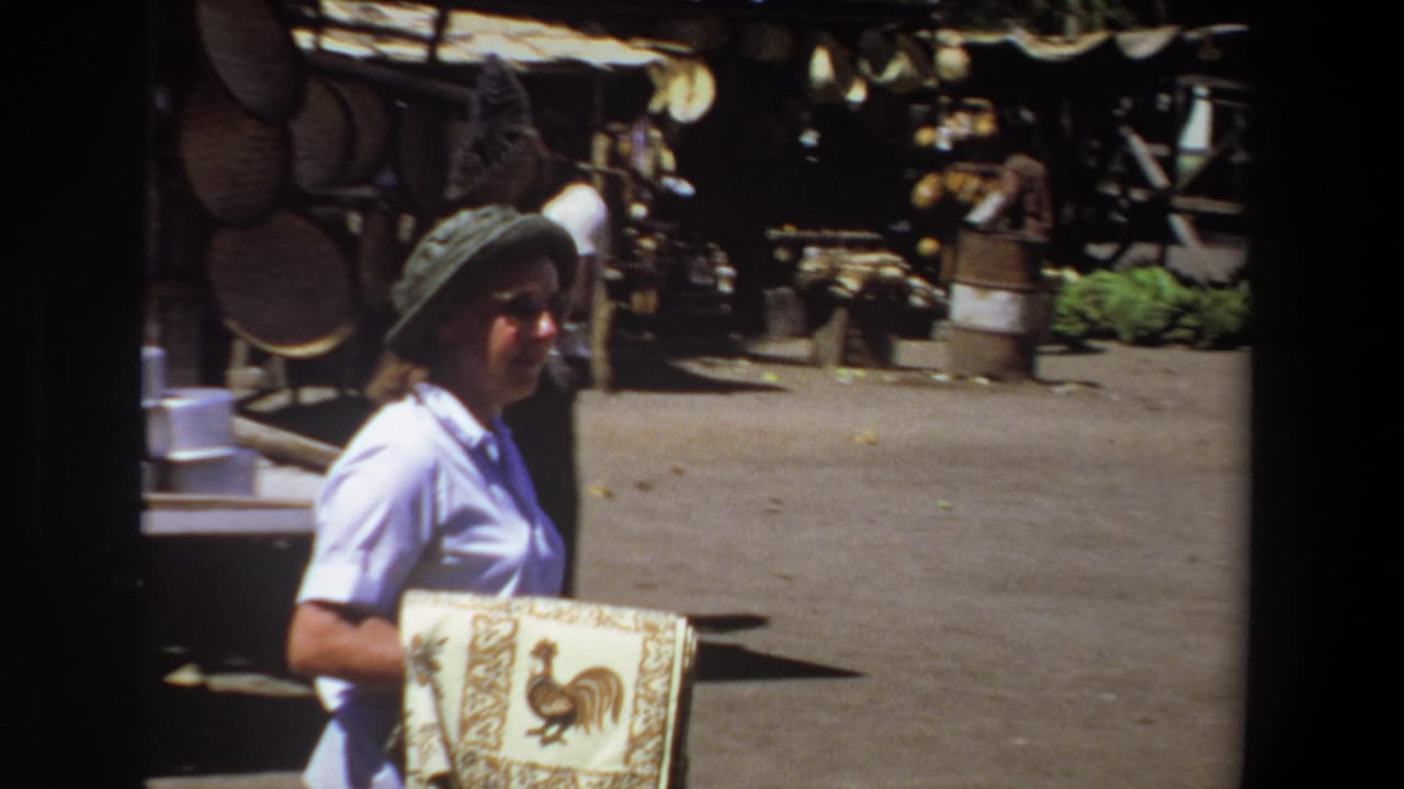 Woman with a hat and cloth walks across an open market area with stalls and people in the background.