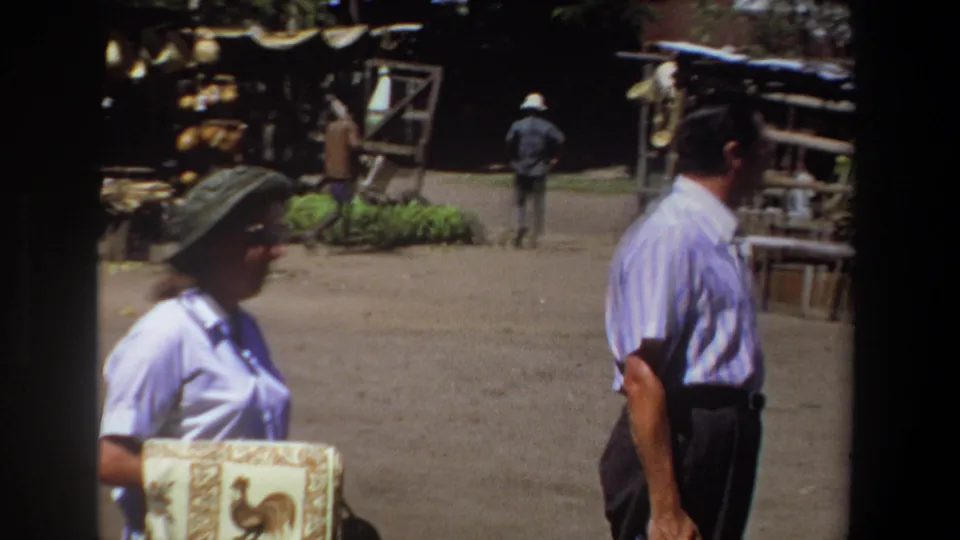 At the Pretoria market, the woman with the rooster-patterned cloth passes a man in a striped shirt as other people walk near the stalls.