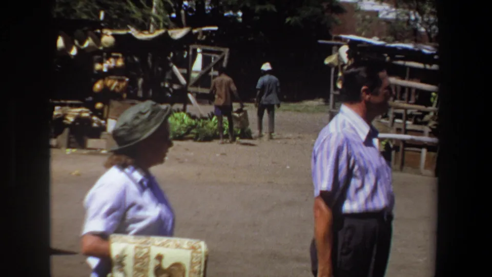 The closing frame shows the woman and a man in a striped shirt in the foreground, with Pretoria market stalls, hanging goods, and pedestrians beyond.