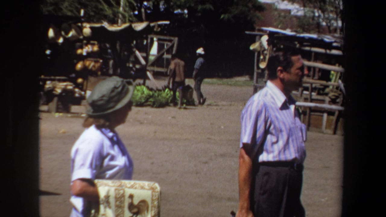 In 1969 Pretoria, a man in a striped shirt and a woman in a brimmed hat pass outdoor market stalls on a dusty street.