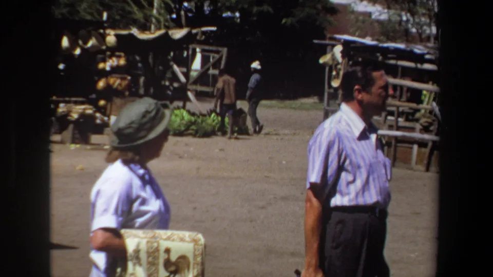 In 1969 Pretoria, a man in a striped shirt and a woman in a brimmed hat pass outdoor market stalls on a dusty street.