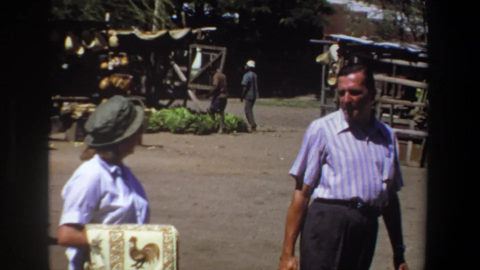 In the Pretoria market scene, the man turns toward the camera while the woman and shaded vendor stalls remain nearby.