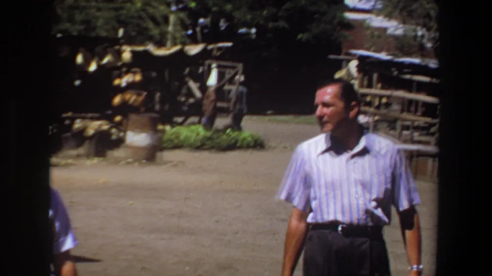 The man continues through the 1969 Pretoria marketplace, with produce displays, shaded structures, and pedestrians in the background.