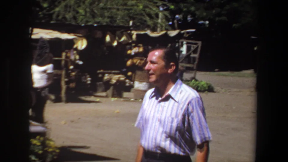 In the closing frame, the striped-shirt man walks beside a shaded stall with hanging goods and produce in the Pretoria market.