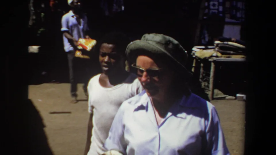 1969 Pretoria market view showing a green-hatted traveler and a local man moving past tables, textiles, and shaded stalls.