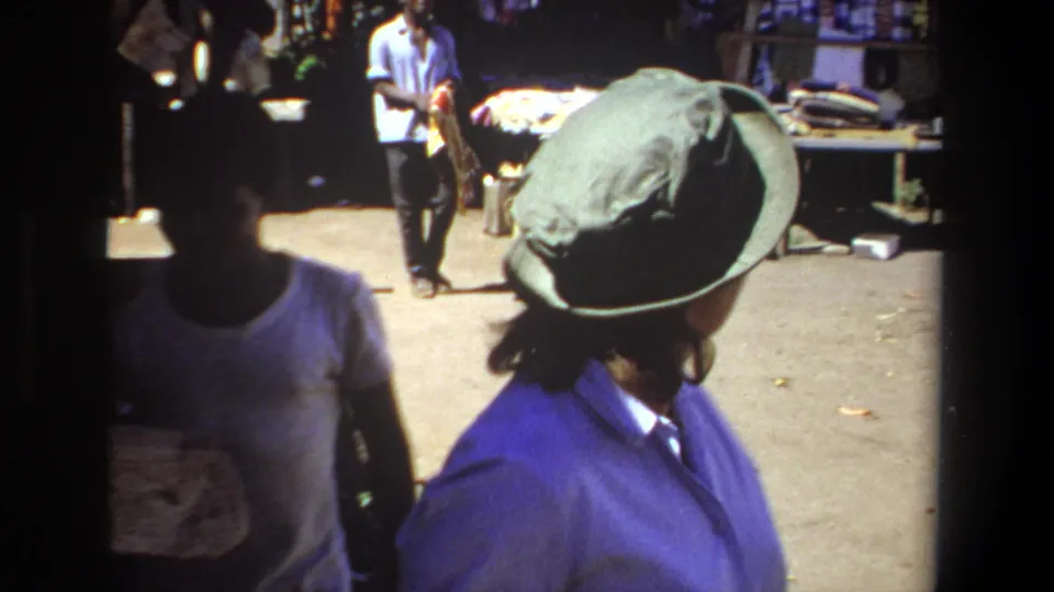 Pretoria market frame with a green-hatted traveler turning near a shaded passerby, vendor tables, and sunlit goods.