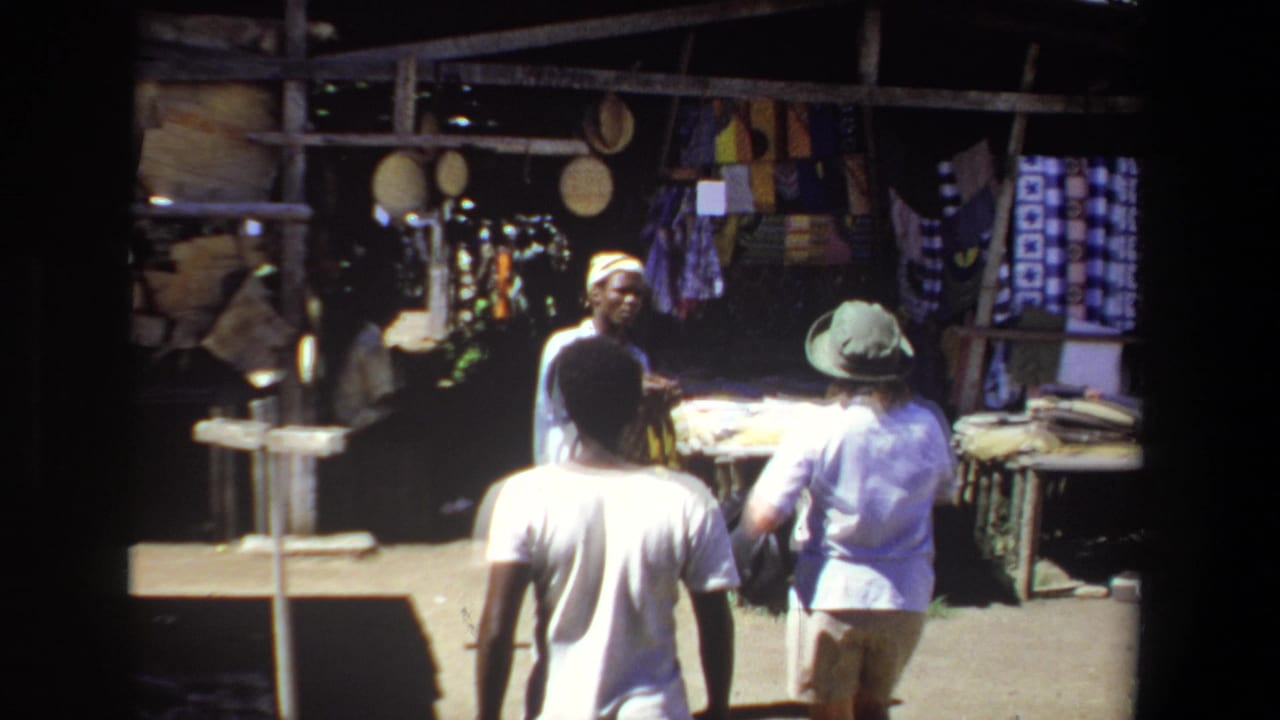 1969 Pretoria home-movie frame showing an outdoor market stall with textiles, a vendor, and visitors browsing beneath a wooden shelter.