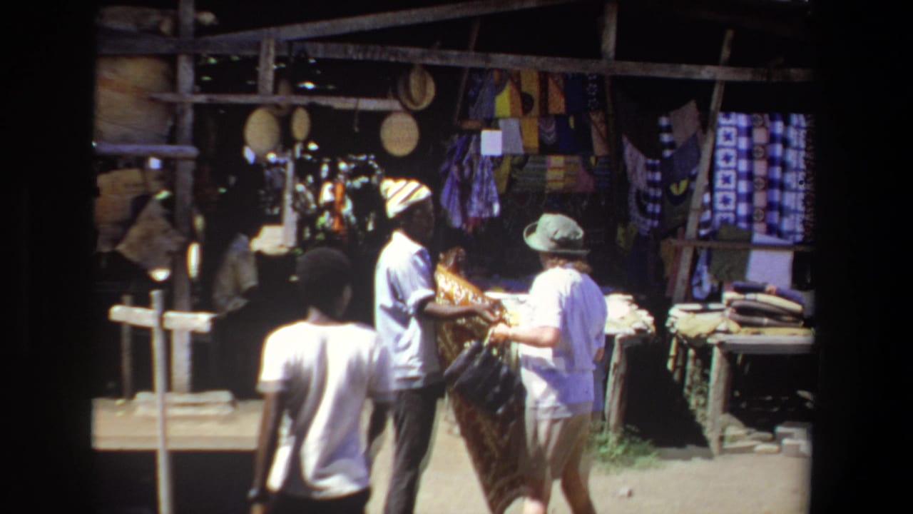 1969 Pretoria market scene with a vendor showing patterned fabric to a visitor in a brimmed hat near hanging textiles and goods.