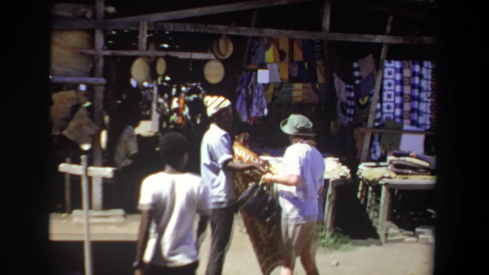 1969 Pretoria market scene with a vendor showing patterned fabric to a visitor in a brimmed hat near hanging textiles and goods.