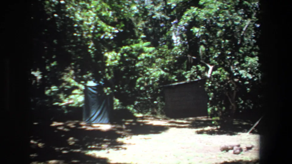 1969 Pretoria-area 8mm frame showing a simple forest campsite with a blue tarp shelter, wooden screen and shaded dirt ground.