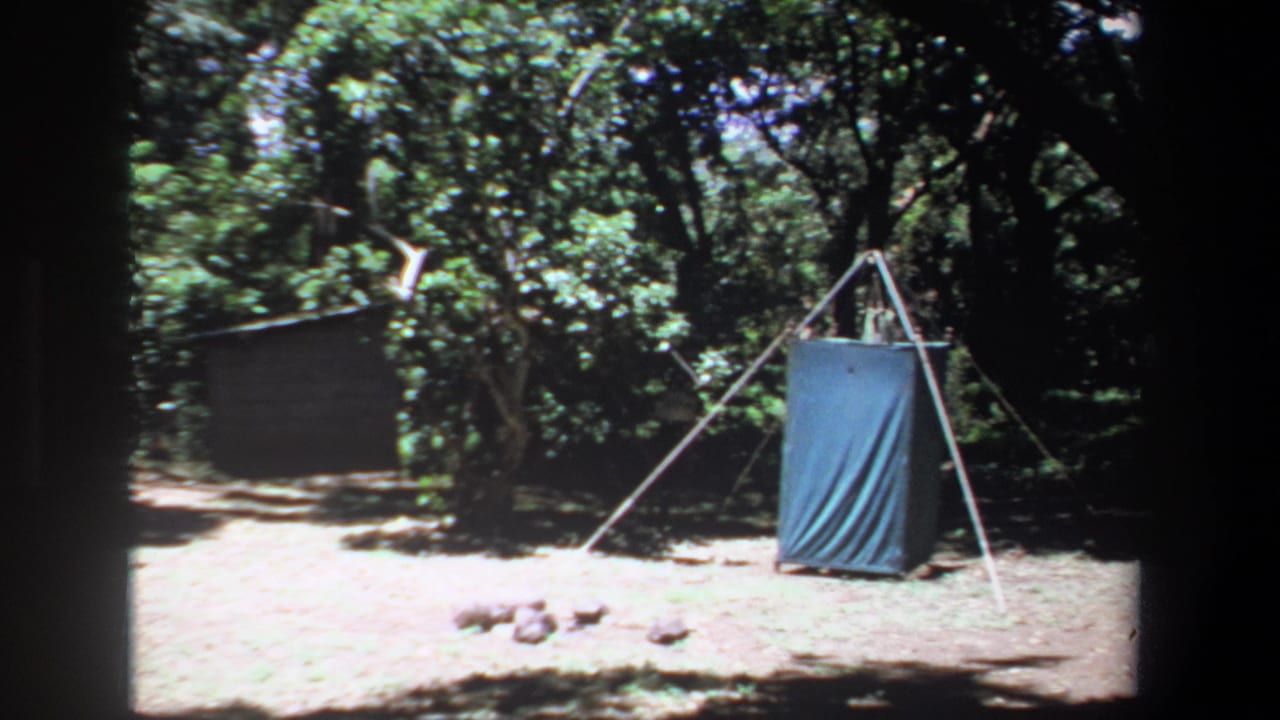 1969 Pretoria-area camping view with a blue tarp shelter on a pole frame, dense trees and stones on the ground nearby.