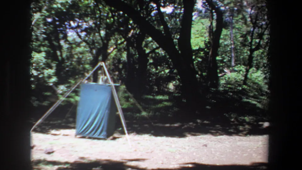 1969 Pretoria-area 8mm frame with the tarp shelter closer in view, surrounded by tree trunks, shade and open dirt ground.