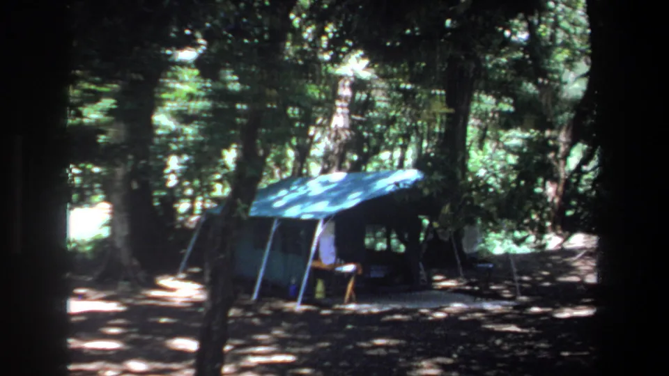 1969 Pretoria home-movie frame showing a blue tent set among trees, with chairs and heavy shade around it.