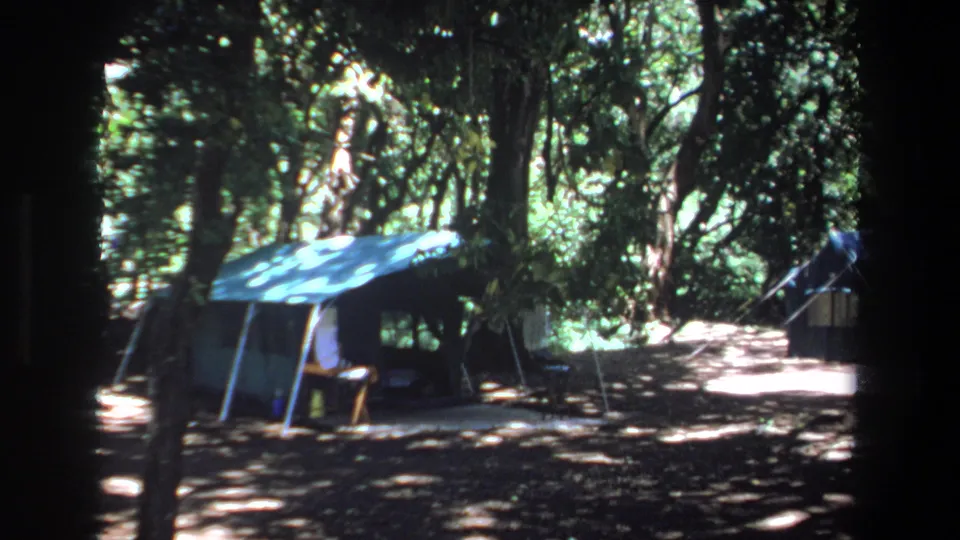 1969 Pretoria footage holds on a blue tent in dense woods, with possible people or belongings in the shadowed shelter.