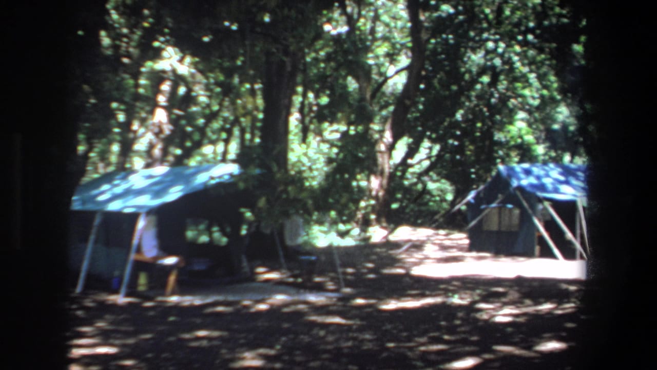 1969 Pretoria home-movie frame showing a shaded campsite with a blue canvas tent, small shelter, and dense trees.