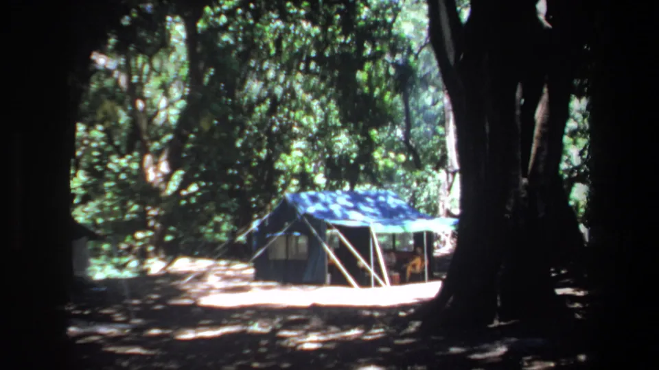 1969 Pretoria camping frame showing a blue tent in a tree-lined clearing with strong shade and dappled light.